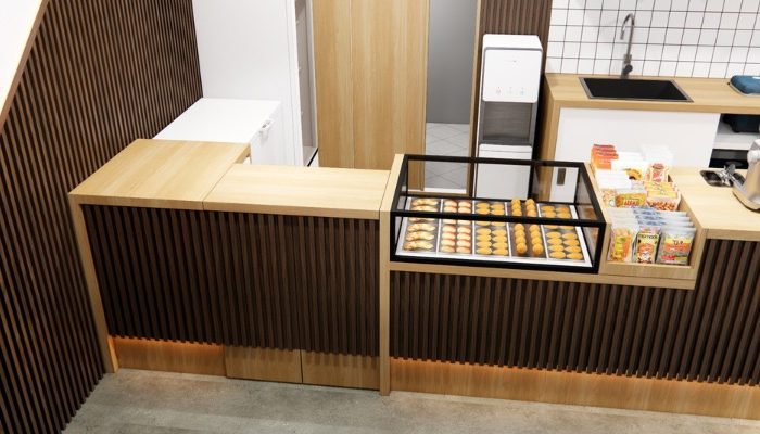 The interior of Cafe Difabis features a modern and inclusive design, incorporating wood elements and warm lighting. The ergonomic barista area is designed for accessibility, showcasing a professional coffee machine, open storage shelves, and a well-organized food display.