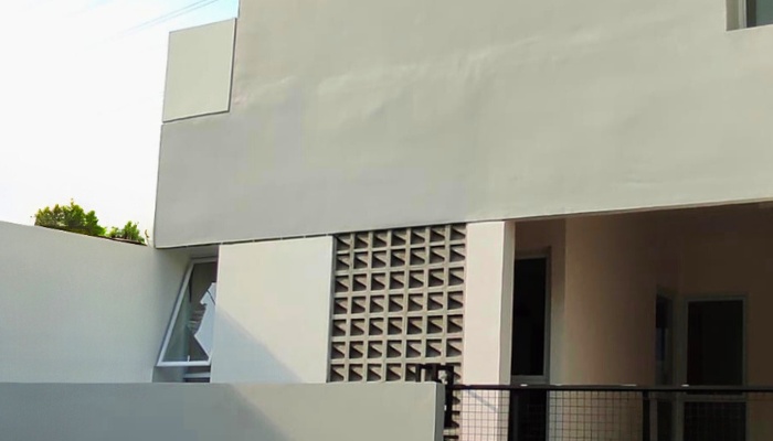 The main façade and interior of WU House in North Jakarta, featuring a tropical modern design with exposed concrete materials, natural wood, and large glass openings that optimize natural lighting and air circulation.