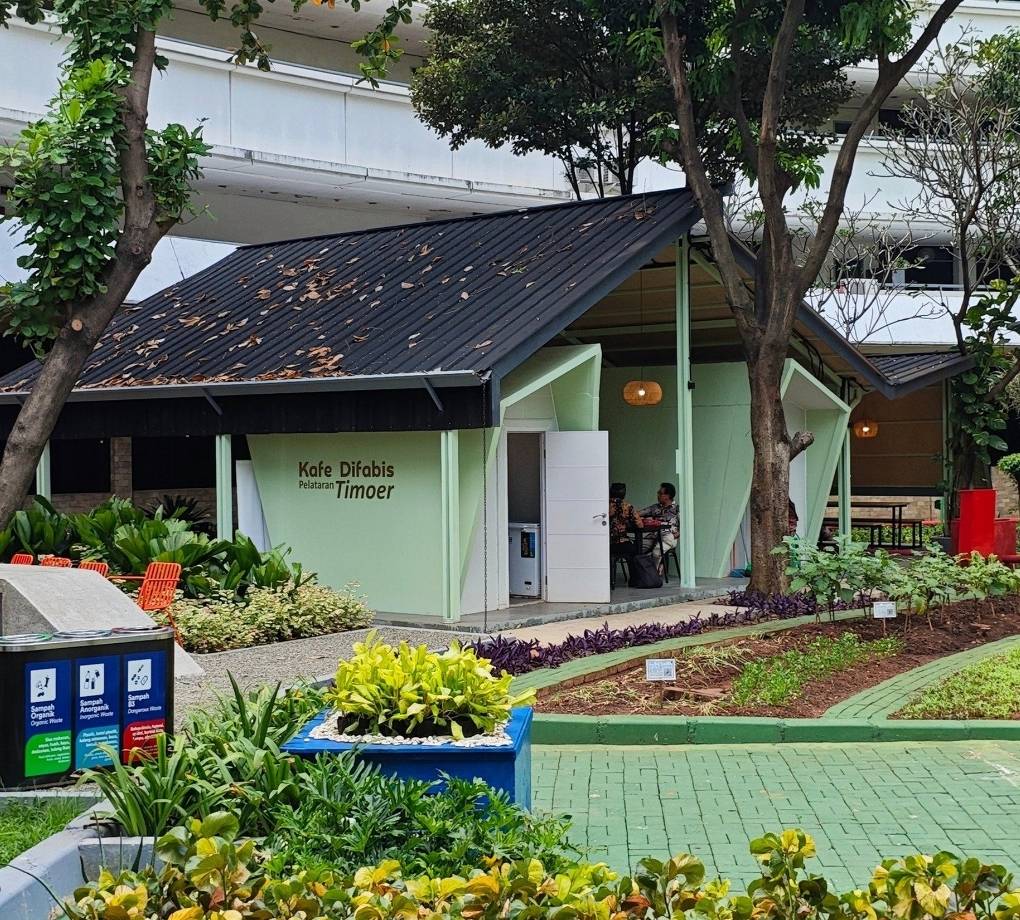 Tampak bangunan Café Difabis Coffee & Bakery karya YPA Architecture Studio di kawasan Kantor Walikota Jakarta Timur dengan atap pelana asimetris, façade hijau pastel, dan area terbuka yang ramah bagi semua pengguna.