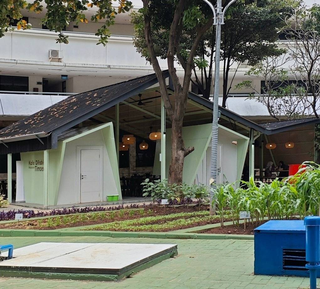 Tampak bangunan Café Difabis Coffee & Bakery karya YPA Architecture Studio di kawasan Kantor Walikota Jakarta Timur dengan atap pelana asimetris, façade hijau pastel, dan area terbuka yang ramah bagi semua pengguna.